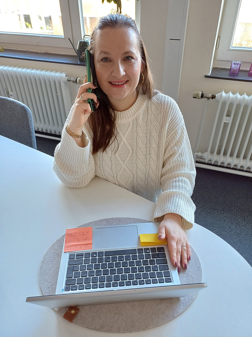 Frau mit weißem Pullover telefoniert und arbeitet am Laptop im Büro, Notizzettel auf dem Gerät, freundliches Lächeln, helle Arbeitsumgebung.
