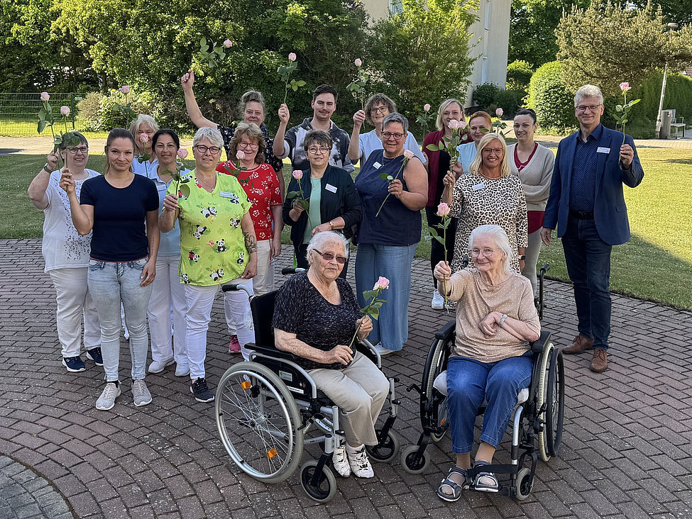 Pflegekräfte, Mitarbeitende und Seniorinnen im Rollstuhl mit rosa Rosen