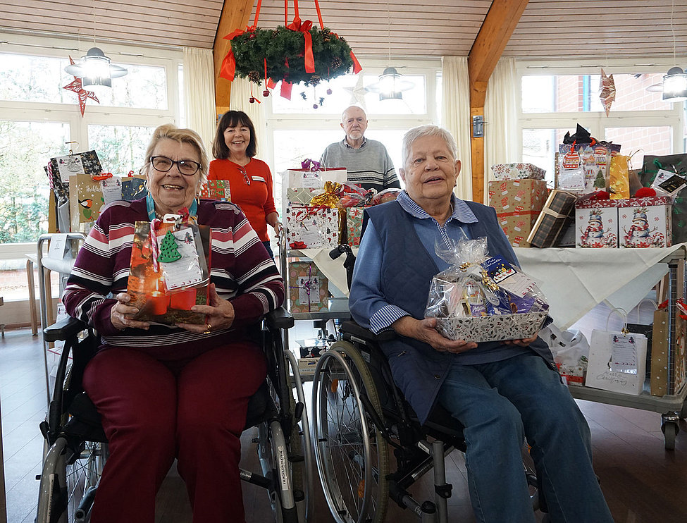 Zwei Bewohnerinnen im Rollstuhl halten Geschenkkörbe vor vielen verpackten Präsenten