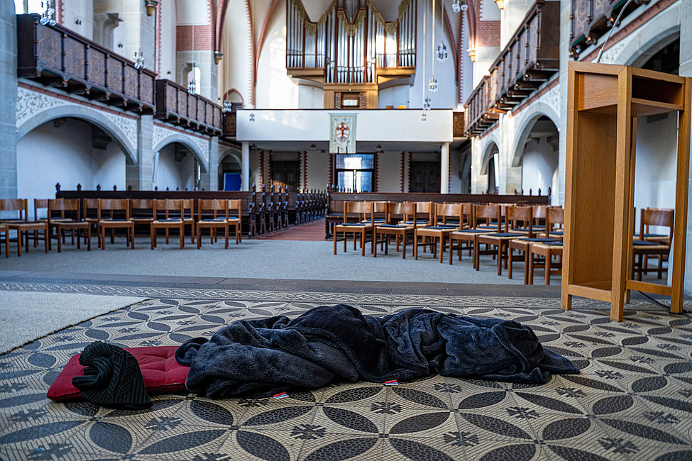 Eine zusammengerollte Decke und ein Kissen liegen auf dem Boden einer Kirche, im Hintergrund leere Stühle und eine Orgel. Die Kirche ist menschenleer.