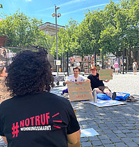 Person mit Shirt „#NOTRUF WOHUNGSMARKT“ im Vordergrund, im Hintergrund zwei Personen mit Protestschildern auf einer Decke auf dem gepflasterten Platz.