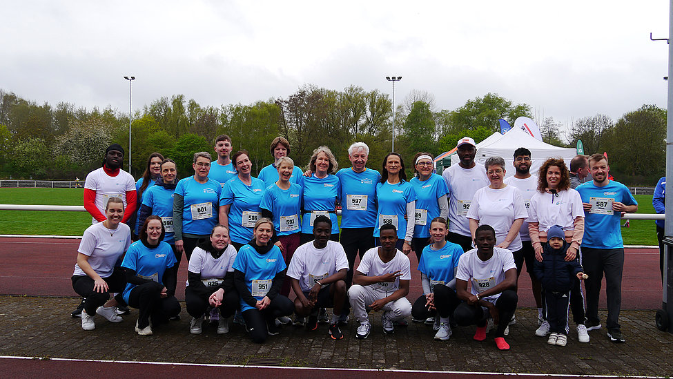 Sportliches Teamfoto: Eine große, gemischte Gruppe strahlt gemeinsam bei einem Event auf der Laufbahn. Alle tragen Wettkampfkleidung und Startnummern.