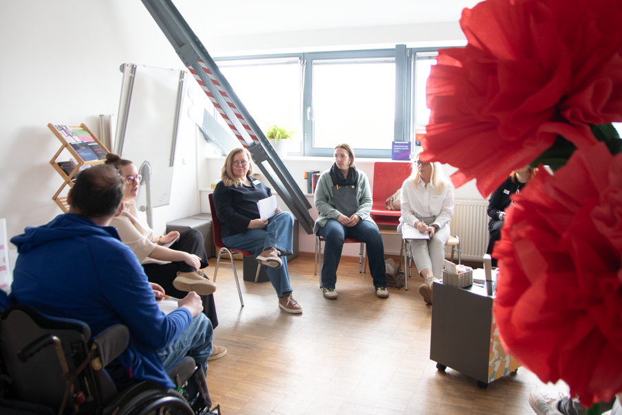 Mehrere Menschen sitzen in einem hellen Raum im Stuhlkreis zusammen. Große, rote Papierblumen befinden sich prominent im Vordergrund.