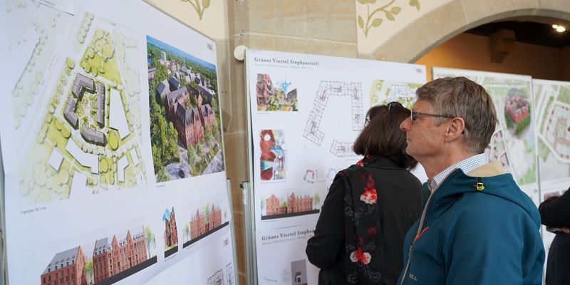 Personen betrachten Ausstellungstafeln mit Architekturplänen und Gebäudevisualisierungen