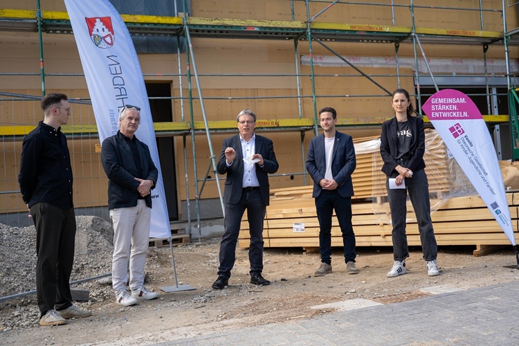 Fünf Personen vor einer Baustelle; eine spricht, zwei Banner stehen links und rechts, Baumaterial und Gerüst im Hintergrund.
