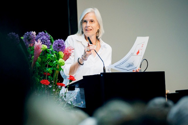 Ältere Frau hält Dokumente am Rednerpult, spricht ins Mikrofon; bunte Blumenarrangements rechts, Publikum undeutlich im Vordergrund.